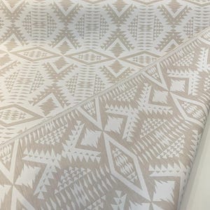 May include: A folded textile with a repeating geometric pattern in beige and white. The design features diamond, triangle, and rectangular shapes, creating a textured, tribal-inspired look. The fabric is likely cotton or linen.