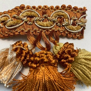 May include: A close-up of a brown, gold, and white tassel trim with a repeating pattern of woven circles. The trim has a fringe of tassels in brown, gold, and white.