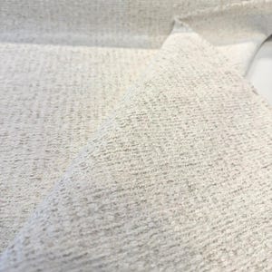 Puede incluir: Primer plano de una tela texturizada en un color blanco roto neutro. El material tiene un aspecto tejido con una superficie ligeramente rugosa. La tela está doblada, revelando su grosor y la dirección del tejido.