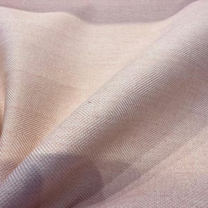 May include: Close-up of a soft, woven fabric in shades of light pink and peach. The fabric has a subtle herringbone pattern, with diagonal lines creating a textured appearance. The folds and shadows add depth to the textile.