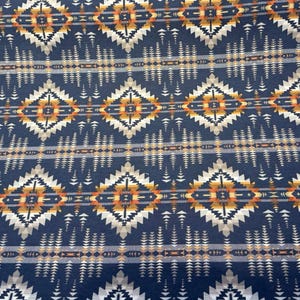 May include: Close-up of a navy blue fabric with a geometric pattern. The design features diamond shapes in white, tan, and orange, along with rows of small triangles and lines. This textile is suitable for various craft projects.