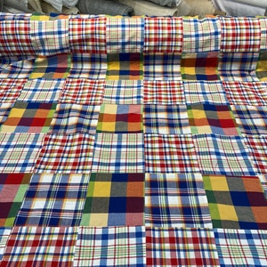 May include: A patchwork fabric with a variety of plaid patterns in red, blue, yellow, and green. The fabric is folded and displayed on a table.