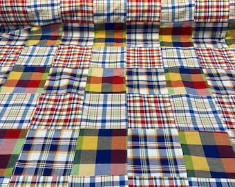 May include: A patchwork fabric featuring a variety of plaid patterns in red, blue, yellow, and green. The fabric is folded and displayed on a table.