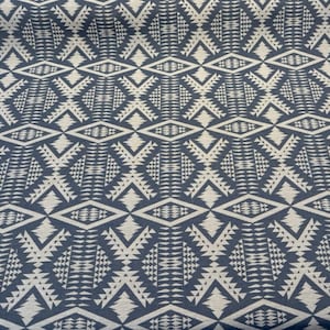 May include: Close-up of a fabric with a repeating geometric pattern. The design features diamond and triangle shapes in a light beige color against a dark blue background. The pattern is symmetrical and covers the entire surface.