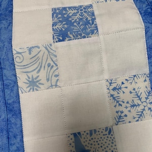May include: A white table runner with a blue border and a patchwork design of blue and white fabric squares. The squares feature various snowflake and floral patterns.