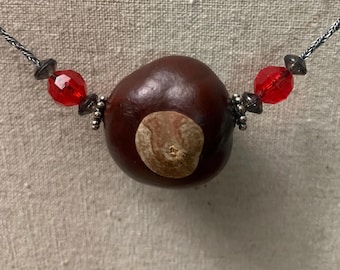 May include: A necklace with a brown chestnut as the centerpiece, surrounded by red glass beads and silver accents.