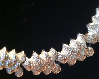 May include: A silver-toned necklace with a textured, scaly design. The necklace is made of linked, overlapping pieces that create a wave-like pattern.