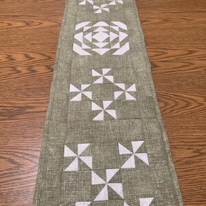 May include: A green table runner with a white geometric pattern. The pattern is made up of pinwheels and squares.