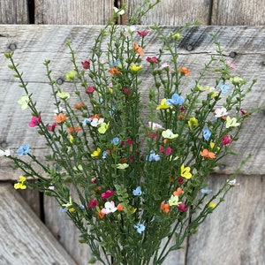 May include: A bouquet of artificial flowers with small, colorful blooms in shades of blue, yellow, pink, and orange. The flowers are arranged on green stems and are held in a hand.