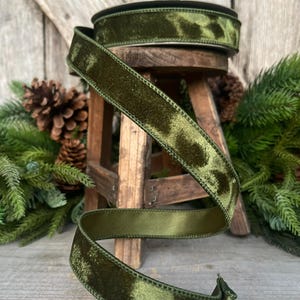 May include: Olive green velvet ribbon, partially rolled, with a satin backing. The ribbon is displayed on a small wooden stool, with pinecones and evergreen branches in the background. The ribbon has a textured, spotted pattern.