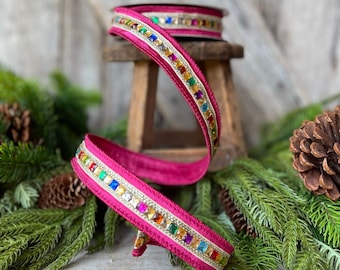 May include: A roll of fuchsia ribbon with a gold border and colorful jewel accents. The ribbon is displayed on a small wooden stool, surrounded by evergreen branches and pinecones, suggesting a holiday theme.