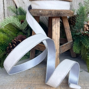 May include: A spool of white velvet ribbon with a silver satin backing and silver trim sits on a small wooden stool. The ribbon is unrolled, displaying its texture and sheen. Pine needles and pine cones are in the background, suggesting a holiday theme.
