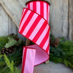 May include: A spool of red and pink striped ribbon. The ribbon is a wide, decorative style, with a bold pattern of vertical stripes. The ribbon is displayed on a wooden surface with greenery in the foreground and background.