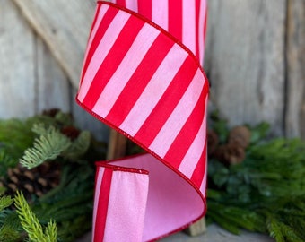 May include: A spool of red and pink striped ribbon. The ribbon is a wide, decorative style, with a bold pattern of vertical stripes. The ribbon is displayed on a wooden surface with greenery in the foreground and background.