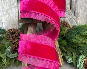 May include: A spool of vibrant fuchsia velvet ribbon with delicate ruffled edges. The ribbon is displayed on a wooden surface with pinecones and evergreen branches in the background, suggesting a holiday or craft theme.
