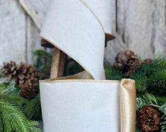 May include: Two spools of white ribbon with gold trim. The ribbon is thick and has a soft, velvety texture. The spools are displayed on a wooden stand, surrounded by pine needles and pine cones, suggesting a holiday theme.