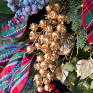 May include: A decorative holiday wreath arrangement featuring green pine needles, large dark green leaves, and a plaid ribbon in red, green, and blue. The display includes clusters of metallic gold berries, frosted blue berries, and bright red berries.