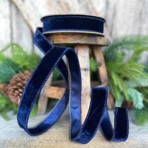 May include: A spool of navy blue velvet ribbon with a satin backing. The ribbon is displayed in a flowing, curved pattern on a wooden surface. The ribbon is approximately 1 inch wide.