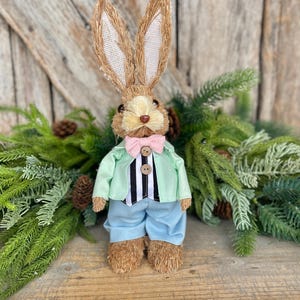 May include: A decorative Easter bunny figurine with a straw-like body, wearing a light green jacket, blue pants, and a pink bow tie. The bunny has long, upright ears and is standing on a wooden surface, surrounded by greenery.