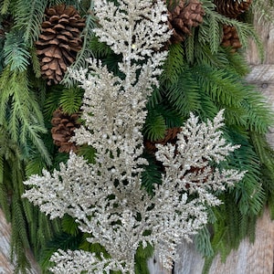 May include: A silver glittered decorative branch with a natural evergreen and pinecone wreath. The branch has a detailed, leafy design. The wreath is made of green pine needles and brown pinecones, set against a rustic wooden background.