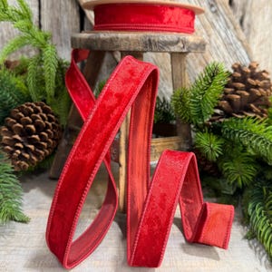 May include: A spool of vibrant red velvet ribbon with stitched edges, draped over a small wooden stool. The ribbon features a soft, textured surface and a deep crimson hue, showcasing a continuous length that cascades down from the wooden spool.