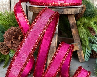 May include: A roll of vibrant magenta ribbon with a shimmering red trim. The ribbon is displayed on a small wooden stool, with a portion unrolled and curled. Pine needles and a pine cone are in the background.