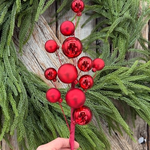 May include: A decorative floral pick featuring multiple round red ornaments of varying sizes attached to thin stems. The ornaments have a mix of glossy and matte finishes. The pick is held against a background of green pine needles.