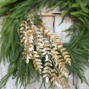 May include: A green wreath with a circular shape, adorned with gold-colored leaf accents. The wreath is made of lush, green foliage, and the gold leaves add a touch of elegance. The wreath is displayed against a rustic wooden background.