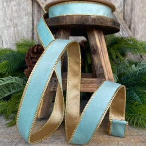 May include: A spool of light blue ribbon featuring a smooth satin finish on one side and a metallic gold edge. The ribbon is draped in loops, showcasing the contrasting gold underside and the shimmering light blue top surface.
