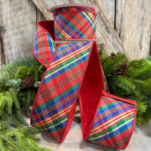 May include: A spool of wired ribbon featuring a vibrant plaid pattern in red, green, blue, and gold. The fabric has a distinct woven texture with thin metallic gold stripes running diagonally across the surface and solid red finished edges.
