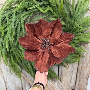 May include: A large, deep brown velvet poinsettia flower with prominent, textured petals and a dark, beaded center. The artificial bloom features visible veining on each petal and is positioned against a backdrop of green pine needles.