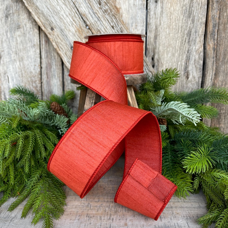 May include: A roll of wide, textured, red ribbon with a matching red trim. The ribbon is displayed with a length of the ribbon unrolled, showing its texture and color. The ribbon is placed on a small wooden stand, surrounded by evergreen branches.