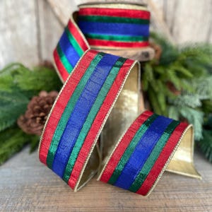 May include: A festive ribbon with red, green, and blue stripes, edged with gold. The ribbon is partially rolled, revealing a spool, and is set against a backdrop of greenery and a wooden surface. Ideal for holiday decorations.