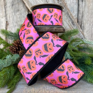 May include: A roll of decorative ribbon featuring a repeating pattern of orange jack-o'-lantern faces on a pink background. The ribbon has black edges and is displayed on a wooden spool, perfect for Halloween crafts.