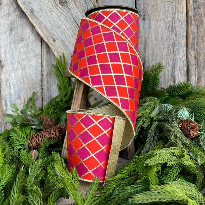 May include: A large roll of decorative ribbon with a repeating diamond pattern in red and fuchsia, accented with gold lines. The ribbon is displayed on a wooden stand, surrounded by evergreen branches and pinecones.