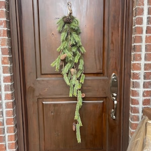 May include: A long, green garland with pine needles and brown pine cones hangs on a wooden door.