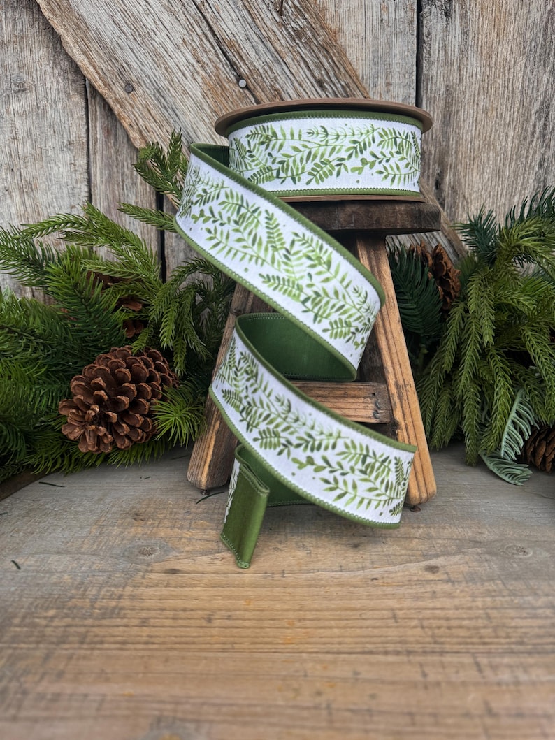 May include: A spool of white ribbon featuring a repeating green fern leaf pattern. The ribbon has a solid green edge and is draped in a spiral shape, showcasing the botanical print against the white fabric background with a finished texture.