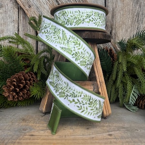 May include: A spool of wired ribbon featuring a white background with a repeating green fern leaf pattern. The ribbon has a solid green underside and finished edges, shown spiraling downward from a wooden spool base with a textured surface.