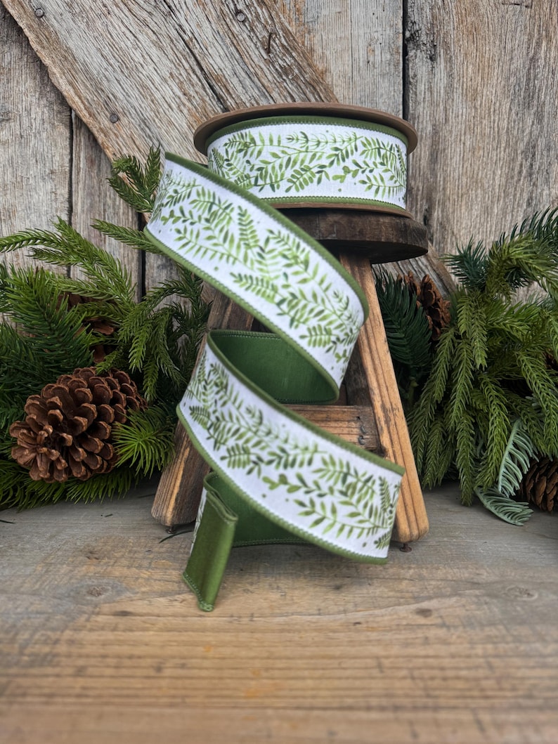 May include: A spool of wired ribbon featuring a white background with a repeating green fern leaf pattern. The ribbon has a solid green underside and finished edges, shown spiraling downward from a wooden spool base with a textured surface.
