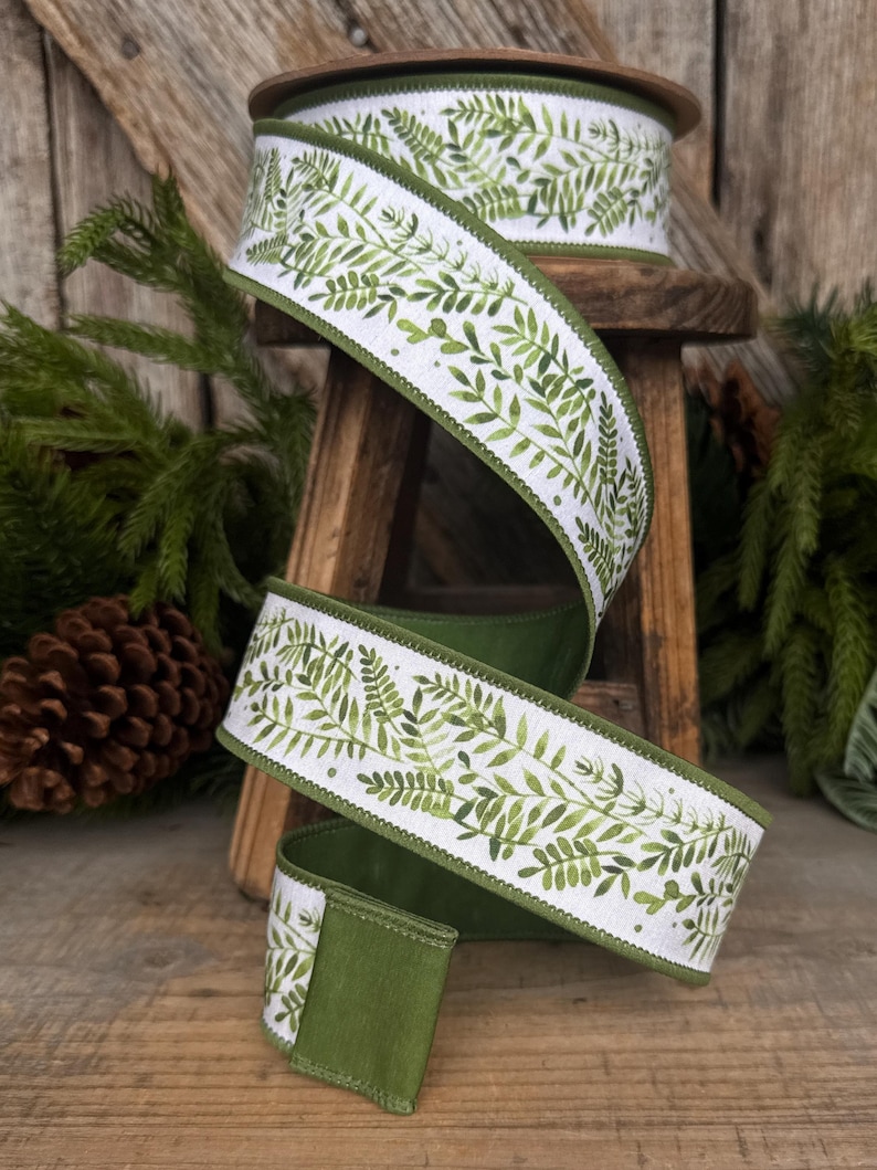May include: A spool of white ribbon featuring a repeating green watercolor fern leaf pattern. The ribbon has a solid green fabric backing and finished stitched edges, shown spiraling downward from a wooden spool.