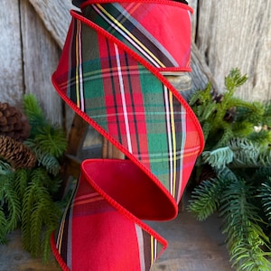 May include: A spool of red and plaid ribbon with a red border. The plaid pattern includes green, black, white, and yellow stripes. The ribbon is displayed against a rustic wooden background with pine needles.