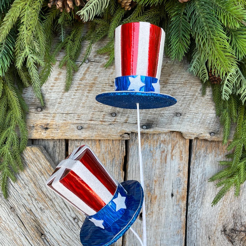 May include: Two red, white, and blue glitter top hats with stars on the band. The hats are on white sticks and are in front of a weathered wooden background.