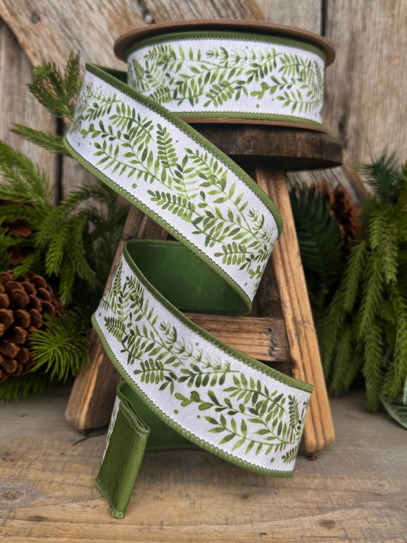 May include: A roll of white wired ribbon featuring a repeating green fern leaf pattern. The ribbon has solid green finished edges and is coiled in a spiral shape, displaying the botanical print clearly against the white fabric background.