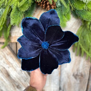 May include: A large, dark blue velvet flower with glittered edges and center. The flower is held in a hand, with a gold watch visible. Green pine needles and a pinecone are in the background. This decorative flower is perfect for holiday decor.