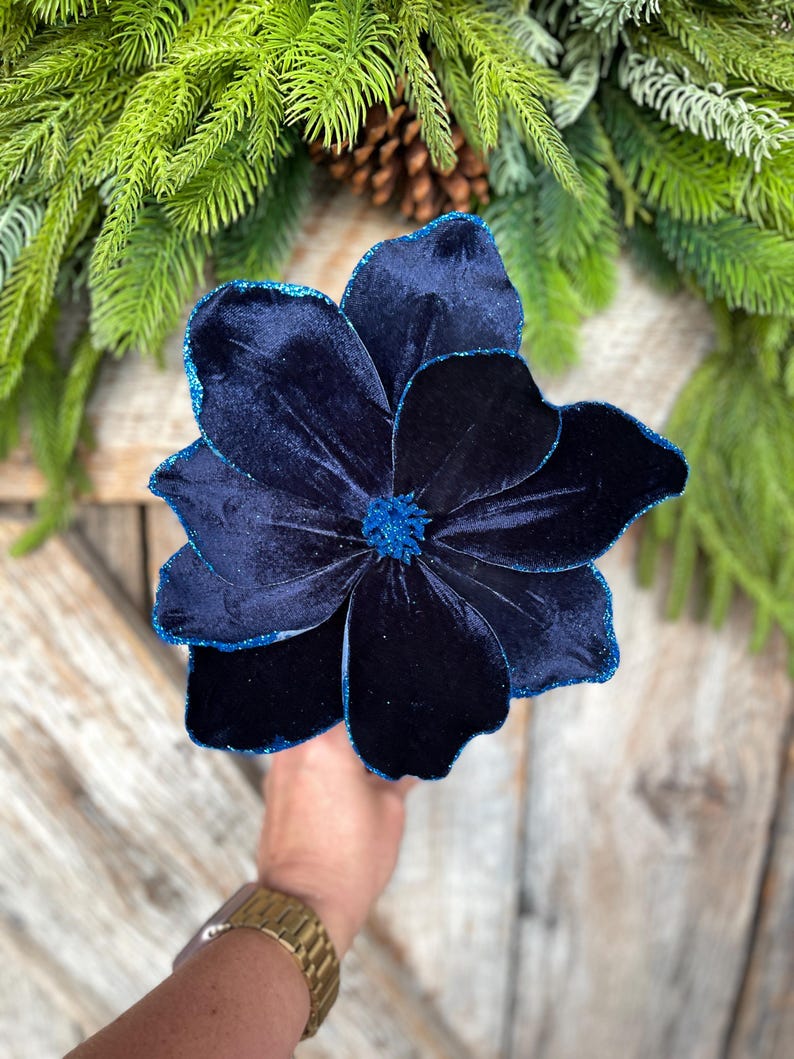 May include: A large, dark blue velvet flower with glittered edges and center. The flower is held in a hand, with a gold watch visible. Green pine needles and a pinecone are in the background. This decorative flower is perfect for holiday decor.