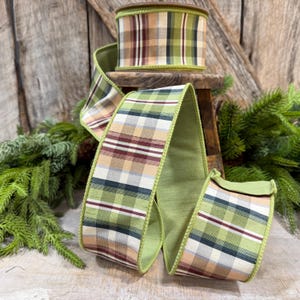 May include: A roll of wired ribbon featuring a plaid pattern in shades of olive green, tan, cream, and dark maroon. The ribbon has a solid olive green backing and a textured fabric finish, displayed in a coiled loop with visible wire edges.