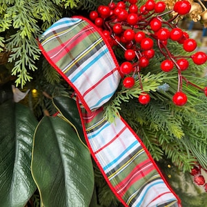 May include: A decorative ribbon featuring a plaid pattern with white, green, red, and light blue stripes, finished with a solid red fabric edge. The ribbon is intertwined with clusters of bright red berries and green pine needles.