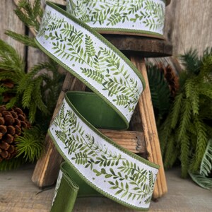 May include: A spool of white ribbon featuring a repeating green watercolor fern leaf pattern. The ribbon has a solid green edge and a wired border, shown unwound in a spiral shape to display the botanical print and the texture of the fabric.