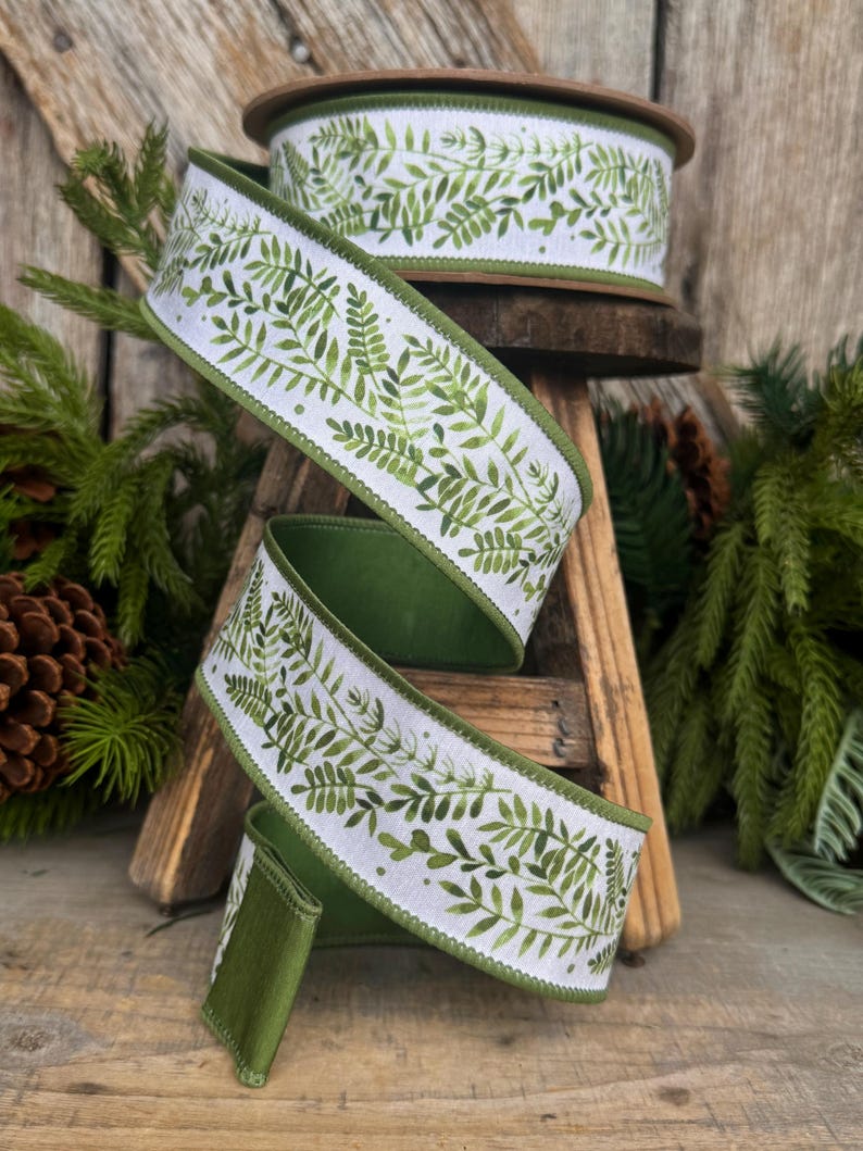 May include: A spool of white ribbon featuring a repeating green watercolor fern leaf pattern. The ribbon has a solid green edge and a wired border, shown unwound in a spiral shape to display the botanical print and the texture of the fabric.