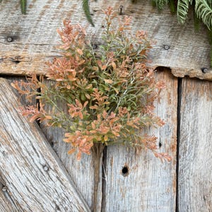 May include: A bundle of artificial greenery with small, pointed leaves in shades of green and orange. The faux plant has a brown stem and is set against a weathered wooden background, suggesting a rustic or farmhouse aesthetic.